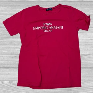 Röd t-shirt från Emporio Armani - Snygg röd t-shirt från Emporio Armani med deras ikoniska logga och texten 'Milan' på framsidan. Perfekt för en stilren och avslappnad look. Tillverkad i mjukt material för extra komfort.