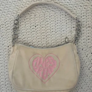 Beige axelväska med hjärtdetalj - Säljer en beige axelväska med en söt rosa hjärtdetalj och texten 'Angel Dream'. Väskan har en dragkedja och en axelrem med silverfärgade kedjedetaljer. Perfekt för att ge en touch av stil till din outfit.