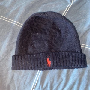 Svart stickad mössa från Polo Ralph Lauren - Snygg svart stickad mössa från Polo Ralph Lauren med röd broderad logotyp framtill. Perfekt för kyliga dagar med sin ribbade kant som ger extra värme och stil.    Inga håll. 