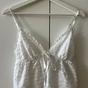 Vit topp - Supersöt vit linnetopp med justerbara smala axelband och v-ringad hals. Toppen har en snygg strukturerad yta och ett knyt framtill som ger extra detalj. Perfekt för dig som gillar en luftig och fräsch look under varma dagar.