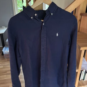 Mörkblå skjorta från Ralph Lauren - Snygg mörkblå skjorta från Ralph Lauren i slim fit med klassisk krage och knappar framtill. Skjortan har lång ärm och ikoniska Polo-loggan broderad på bröstet. Tillverkad i mjuk bomull som känns skön mot huden. Perfekt för dig som gillar stilrena och tidlösa plagg.