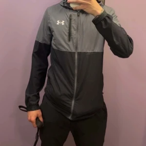 Grå och svart vindjacka Under Armour - ( Kedjan går att dra upp och ner men inte ut till botten ,man måste sätta på den igenom huvudet)Snygg vindjacka från Under Armour i tvåfärgad design med grå upptill och svart nedtill. Jackan har huva, dragkedja framtill och en bröstficka med zip. Perfekt för dig som gillar sportig stil och vill ha något lätt och funktionellt.
