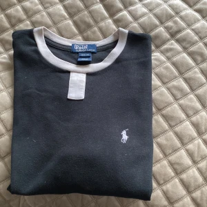 Svart T-shirt från Polo Ralph Lauren - Svart topp från Polo Ralph Lauren med vita kontrastdetaljer. Klassisk broderad logga på bröstet. Tröjan har korta ärmar med vit kant och är gjord i mjuk bomull. Perfekt för dig som gillar stilrena märkesplagg.