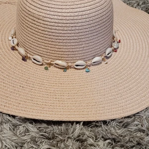 Beige solhatt med snäckor och pärlor - Säljer en stor beige solhatt med bred brätte, dekorerad med snäckor och små färgglada pärlor runt kronan. Hatten har en tunn snörning som kan knytas framtill.
