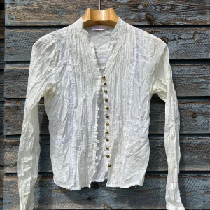 Vit långärmad blus från Zoul - Vit blus från Zoul med lång ärm, dekorativa knappar i guld och snygga spetsdetaljer längs sömmarna. Blusen har en v-ringad hals och tunna vertikala veck framtill som ger en cool vintagekänsla. Perfekt för dig som gillar detaljer och stilren look.