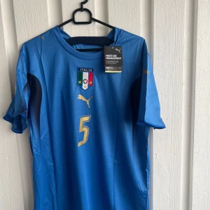 Italien Cannavaro #5 Puma fotbollströja - Snygg blå Italien fotbollströja från Puma med guldiga detaljer, nummer 5 och Cannavaro på ryggen. Tröjan har korta ärmar, broderat landslagsemblem och tryckt Puma-logga. Materialet är lätt och andas, perfekt för match eller träning.