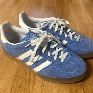 Adidas Gazelles - Snygga Adidas Gazelle sneakers i blå mocka med vita detaljer och klassiska tre ränder på sidan. Skorna har vit skinnpanel vid hälen, vit snörning och genomskinlig gummisula. Perfekt för dig som vill ha en trendig look och vill sticka ut med färg. Skorna kommer även med blåa skosnören (smsa för bild). Tyvärr kommer skorna inte med lådan eftersom jag kunde inte hitta den (förmodligen har den kastats) så de kommer att skickas i en annan skolåda, men de har användts 1 gång. Hör av er vid frågor💗💗💗
