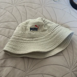 Beige bucket hat från Puma - Snygg beige bucket hat från Puma med broderad logga i blått och rött framtill. 