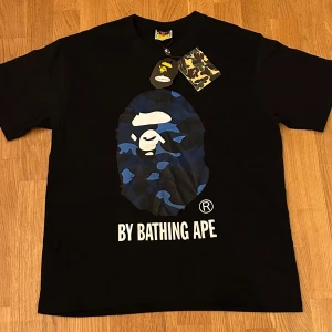 Svart BAPE t-shirt med blå camo print - Svart t-shirt från BAPE med ikoniskt ape-huvud i blått camouflage tryck på bröstet och texten 'BY BATHING APE' under. Rund hals och korta ärmar.  perfekt för streetwear och att sticka ut med cool grafik.