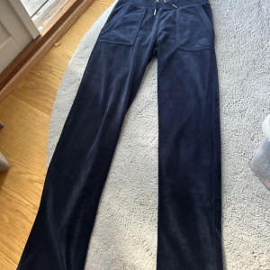 Mörkblå mjukisbyxor Juicy Couture XXS - Snygga mörkblå mjukisbyxor från Juicy Couture i supermjuk velour. Byxorna har snörning i midjan, stora fickor fram och en bakficka med metallknappar. Perfekta för chill eller häng med kompisar. Ikonisk Juicy Couture-logga broderad på fickan.