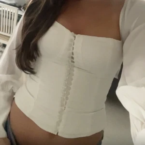 Vit korsettblus med knyt och puffärm - Säljer en vit korsettblus med puffiga långärmar och snygg knäppning framtill. Toppen har öppen rygg med knytband i nacken och sitter tight över midjan. Materialet är lätt och ger en fräsch look, perfekt till jeans eller kjol.