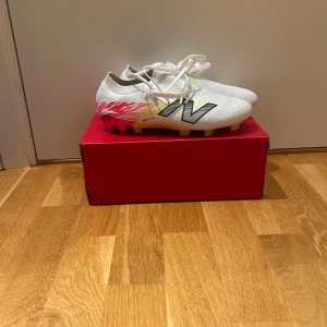 New Balance vita fotbollsskor 42,5 - Säljer ett par grymma New Balance fotbollsskor i storlek 42,5. Skorna är vita med coola detaljer i rosa, gul och svart, och har snörning samt en platt sula med dobbar för bästa grepp på planen. Lätta och snygga med sportig design, perfekta för dig som vill sticka ut. Nu pris 3199 mitt pris 1499 aldrig använda 