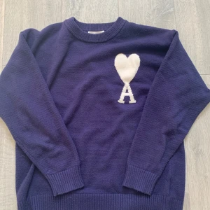 Ami Paris tröja - Säljer en mörkblå stickad tröja från Ami med ett stort vitt hjärta och bokstaven A på bröstet. Tröjan har rund hals, ribbade muddar och lång ärm. Perfekt för dig som gillar stilrena detaljer och vill ha något som sticker ut lite extra. Storleken är M men passar också bra på S. Priset är inte huget i sten
