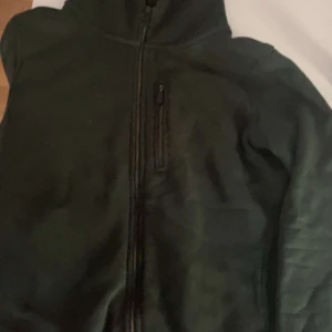 Mörkgrön fleecejacka med dragkedja - Snygg mörkgrön fleecejacka med hel dragkedja framtill och en extra ficka med dragkedja på bröstet. Jackan har en enkel design och är perfekt för kyliga dagar. Materialet är mjukt och bekvämt, och färgen är riktigt stilren.