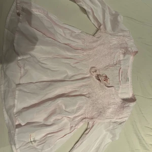 Odd Molly ljusrosa blus med rosett - Supersöt ljusrosa blus från Odd Molly med smock upptill och en fin rosett vid halsen. Blusen har långa ärmar med volangdetaljer och spetskant, samt en luftig och flowy passform. 