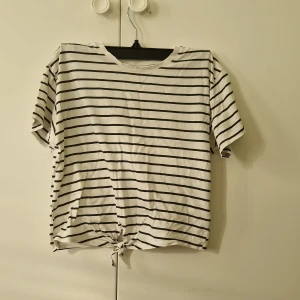 Randig vit t-shirt med knytet där fram från H&M - Vit t-shirt från H&M med svarta ränder och korta ärmar. Den har en knytning längst ner framtill. Storlek 152 för barn