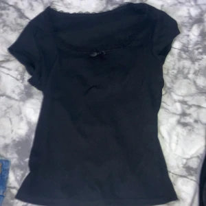 Svart topp med spets och rosett från SHEIN - Svart kortärmad topp från SHEIN i ribbad bomullsmix. Söt spetskant runt halsen och en liten rosett framtill ger en gullig vibe. Passformen är slim och toppen är perfekt att styla med jeans eller kjol. Storlek XS.