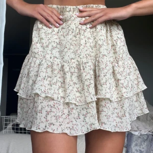 Blommig volangkjol  - Supersöt kort volangkjol i ljusbeige med små blommor i rosa och grönt. Kjolen har två lager volanger och resår i midjan för en bekväm passform. Perfekt för dig som gillar en romantisk och somrig stil.