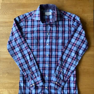 Rutig blå och röd skjorta från Eton - Snygg slim fit skjorta från Eton i storlek S med rutigt mönster i blått och rött. Kragen har en cool detalj med små blommor i rosa och gult mot mörkblå bakgrund. Skjortan har långa ärmar och klassiska knappar framtill. Perfekt för dig som gillar stilrena och unika detaljer.