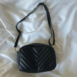 Svart quiltad axelväska avtagbar axelrem - Snygg svart axelväska med quiltad design i sicksackmönster. Väskan har en justerbar axelrem och stängs med dragkedja i metall. Perfekt storlek för mobil, plånbok och nycklar. Stilren look som passar till många outfits