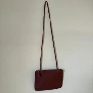 Brun axelväska från Sandqvist - Snygg brun axelväska från Sandqvist i slätt skinn. Väskan har en minimalistisk rektangulär form, silverfärgade metalldetaljer och dragkedja upptill. Perfekt storlek för mobilen och småsaker