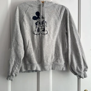 Grå Disney Mickey Mouse hoodie M - Säljer en grå hoodie från Disney med ett stort svart tryck av Mickey Mouse på framsidan. Tröjan har huva med snörning, magficka och ribbade muddar vid ärmslut och nederkant. Perfekt för dig som gillar streetstyle och vill ha något bekvämt och snyggt.