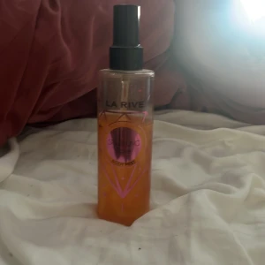 La Rive Sparkling Rose Body Mist - Fräsch body mist från La Rive med doften Sparkling Rose. Flaskan är genomskinlig med rosa detaljer och svart spraykork. Perfekt för dig som gillar lätta och blommiga dofter. Innehållet är orangefärgat och flaskan har en modern, stilren design.