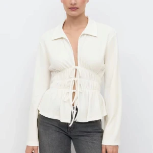 Vit skjorta med snörning och krage - Snygg vit skjorta med krage och markerad midja med snörning framtill. Skjortan har långa ärmar och ett lätt rynkat parti vid midjan som ger en cool siluett. Perfekt för dig som gillar trendiga detaljer och stilren look.
