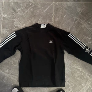 Svart Adidas sweatshirt med vita ränder - Svart sweatshirt från Adidas med klassiska vita ränder längs ärmarna och Adidas-logga på bröstet och ärmen. Tröjan har rund hals och lång ärm, tillverkad i mjuk bomullsmix som är skön mot huden. Perfekt för en sportig och chill stil.