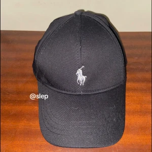Svart keps från Polo Ralph Lauren - Snygg svart keps från Polo Ralph Lauren med klassisk böjd skärm och broderad logga i vitt framtill. Kepsen har en stilren design och är tillverkad i bomull, perfekt för dig som vill ha en clean look. Använts 1 gång!