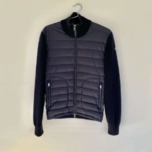 Svart Moncler pufferjacka med stickade ärmar - Snygg svart pufferjacka från Moncler med stickade ärmar och hög krage. Framsidan är quiltad med dunfyllning och har två dragkedjeförsedda fickor. Ärmarna är stickade och har Moncler-logga på vänster arm. Perfekt för dig som gillar exklusiva märken och stilren design.