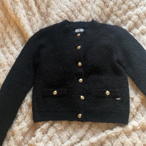 Svart kofta med guldknappar Holly & Whyte - Snygg svart kofta från Holly & Whyte med exklusiva guldknappar framtill och på fickorna. Koftan har en mjuk, stickad känsla och långärmad passform. Perfekt för dig som gillar klassisk stil med en twist. Passar dig som vill ha något både stilrent och coolt.