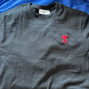 Svart t-shirt från Ami med rött hjärta - Svart t-shirt från Ami med klassisk rund hals och korta ärmar. På bröstet sitter ett broderat rött hjärta med bokstaven A, som ger en snygg och unik detalj. Tillverkad i mjuk bomull som känns skön mot huden. Perfekt för dig som gillar stilrena plagg med en twist.