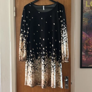 Svart och beige blommig klänning Chiara Point - Supersöt kortklänning från Chiara Point Milano med svart överdel och beige blommigt mönster som tonar ut mot fållen och ärmarna. Långärmad med lätt puff och resår vid ärmslut. Mjuk polyester som faller snyggt och känns skön mot huden.