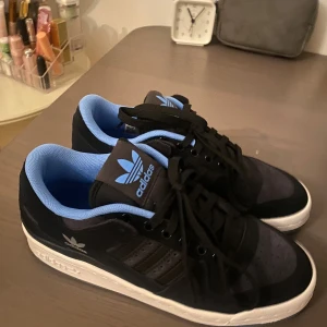 Adidas Forum 84 Low ADV svart/blå - Säljer ett par Adidas Forum 84 Low ADV sneakers i svart och blått med vit sula och genomskinlig undersida. Skorna har klassisk snörning, rund tå och Adidas logga i blått på plösen. Insidan är mjukt vadderad i blått och ovandelen är i syntet och skinn.