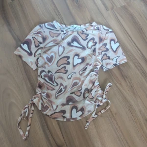 Beige mesh topp med hjärtan FB Sister - Säljer en beige mesh topp från FB Sister i storlek XXS. Toppen har korta ärmar, coolt hjärtmönster i bruna och vita toner samt snygga snörningsdetaljer på sidorna som ger en croppad vibe. Perfekt för dig som gillar statement-plagg med unik design.