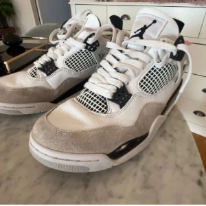 Nike Jordan 4 Retro vit/svart/grå - Nike Jordan 4 Retro sneakers i vit, svart och ljusgrå färg. Skorna har meshpaneler, snörning och detaljer i mocka och skinn. Ikonisk Jumpman-logga på hälen och tungan, samt platt sula och rund tå. Perfekt för dig som vill ha en clean och sportig look.