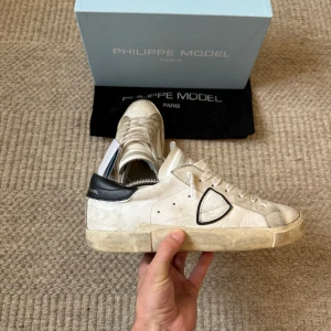 Philippe Model PRSX - Säljer ett par Philippe Model sneakers i vitt med svart hälparti och snygga detaljer på sidan. Skorna har snörning, rund tå och platt sula. Materialet är skinn och mocka, med en cool kontrast mellan den ljusa ovandelen och den svarta hälen. Perfekt för dig som gillar stilrena och trendiga sneakers.