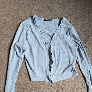 Ljusblå ribbad blus från SHEIN - Supersöt ljusblå blus från SHEIN med ribbat tyg och smala vertikala ränder. Croppad passform, långa ärmar med lätt rynk vid ärmslut och volangkant längs knäppningen framtill. Perfekt för dig som gillar stilrena detaljer och mjuka färger. Nästan aldrig använd