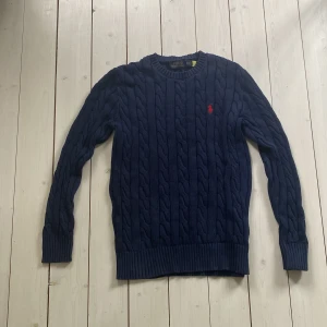 Marinblå kabelstickad tröja Polo Ralph Lauren - Snygg marinblå kabelstickad tröja från Polo Ralph Lauren i storlek XS. Tröjan har ett klassiskt rött broderat logomärke på bröstet, rund hals och ribbade muddar. Perfekt för dig som gillar stilrena och tidlösa plagg med premiumkänsla.