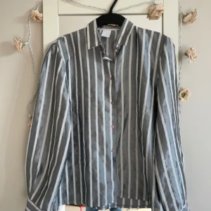 Randig skjorta från Paige i grått - Snygg  vintage skjorta från Paige med vertikala vita och grå ränder. Klassisk krage, långa ärmar och knappar framtill. Skjortan har en normal passform och är gjord i ett lätt, vävt material som känns skönt mot huden. Perfekt till jeans för en chill look.har köpt den från år 1988 .fortfarande fin är den 
