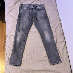 Grå jeans med straight fit - Snygga grå g-raw jeans med tvättad look och klassisk femficksdesign. Modellen har en rak passform som sitter skönt och ger en avslappnad vibe. Jeansen har knapp och dragkedja framtill samt bälteshällor. Perfekt för dig som gillar stilrena och enkla jeans.