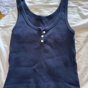 Mörkblå croppad ribbad linnetopp med knappar - Snygg mörkblå ribbad linnetopp med rundad hals och tre vita knappar framtill. Modellen är ärmlös och har en klassisk, slim passform som sitter skönt mot kroppen. Perfekt att styla med jeans eller shorts för en chill look.
