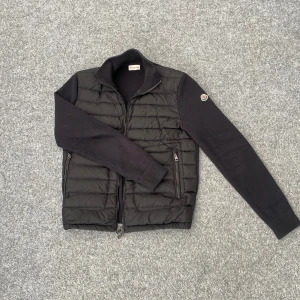 Moncler cardigan  - Svart pufferjacka från Moncler i storlek S med quiltad front och stickade ärmar. Jackan har dragkedja framtill, två sidofickor med dragkedja och Moncler-logga på vänster ärm. Snygg, stilren och perfekt för kyliga dagar.