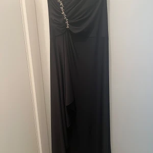 Svart långklänning med pärldetaljer - Elegant svart långklänning med draperad överdel och snygga pärldetaljer längs bröstet. Klänningen har en mjuk, flytande passform och är axelbandslös, perfekt för en extra glam look. Materialet känns lent och har ett fint fall.