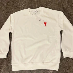 Vit sweatshirt från Ami Paris med rött hjärta - Vit sweatshirt från Ami Paris med klassisk rund hals och lång ärm. På bröstet sitter Ami:s ikoniska röda hjärta med bokstaven A. Tröjan är i mjuk bomull och har ribbade muddar vid ärmslut och nederkant. Perfekt för en clean och stilren look.