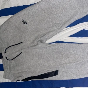 Grå mjukisbyxor från Nike, storlek L - Säljer ett par grå mjukisbyxor från Nike i storlek L. Byxorna har svart snörning i midjan, svart Nike-logga på vänster lår och en svart dragkedja vid ena benet. Tillverkade i mjukt bomullsmaterial, perfekta för chill eller träning.