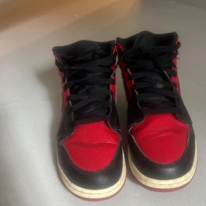 Nike Air Jordan 1 svart/röd strl 39 - Säljer ett par Nike Air Jordan 1 mid banned i klassisk svart och röd färg. Skorna har snörning, rund tå och platt sula. Ovandelen är i skinn med ikoniska swoosh-loggan på sidan och Air Jordan-vingen vid ankeln. Perfekt för dig som gillar streetstyle och sneakers med attityd. Skorna är äkta och köpta för ett tag sen men kommer ej till användning längre