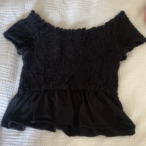 Svart offshoulder topp Gina Tricot XS - Supersöt svart offshoulder topp från Gina Tricot i storlek XS. Toppen har smockad överdel med volangkant nedtill och korta ärmar. Perfekt att styla med jeans eller kjol för en trendig look. Materialet är mjukt och stretchigt, vilket gör den bekväm att bära hela dagen.