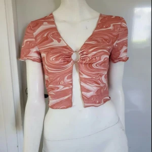 Swirlmönstrad croppad topp med ring - Säljer en kort, swirlmönstrad topp i ljusrosa och beige toner. Toppen har v-ringning, korta ärmar och en metallring framtill som samlar tyget och ger en snygg detalj. Materialet är tunt och stretchigt, perfekt för varma dagar. Passformen är slim och toppen är croppad.